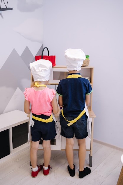 Little biy and girl stand back to camera and play on toy kitchen they dressed in chefs jackets