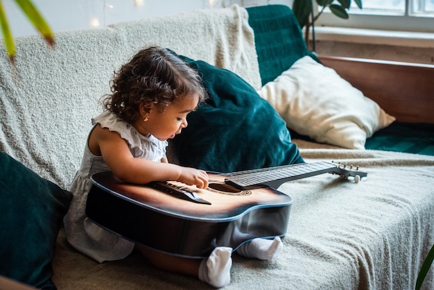 A little black girl in a hat sits on a sofa and plays the guitar baby is playing composes melody and music