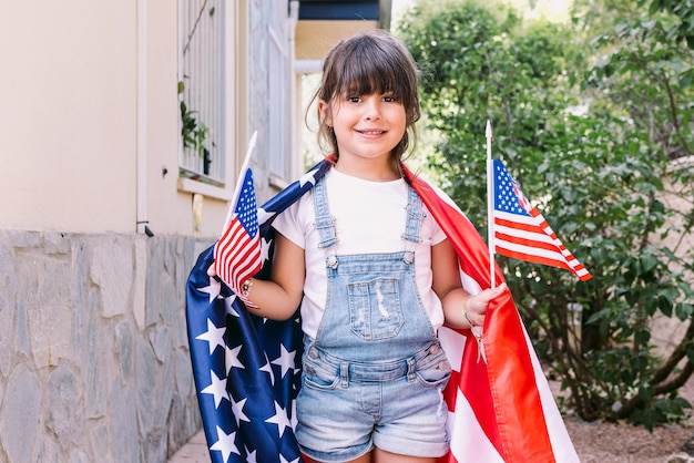 Little blackhaired girl covered with big USA flag holding little American flags in her home garden Concept of celebration independence day 4th of July patriotism holiday and American pride