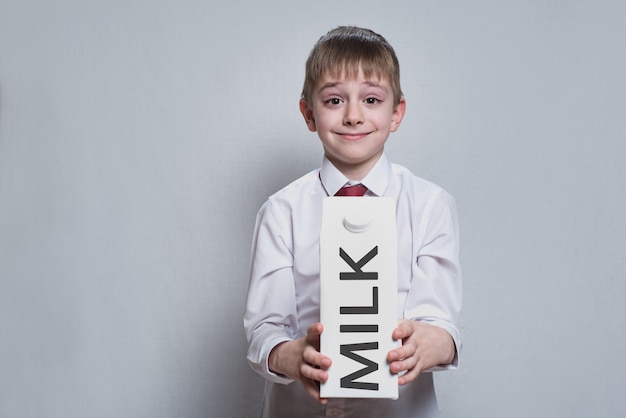 Little blond boy holds and shows a big white carton milk package. White shirt and red tie. Light 