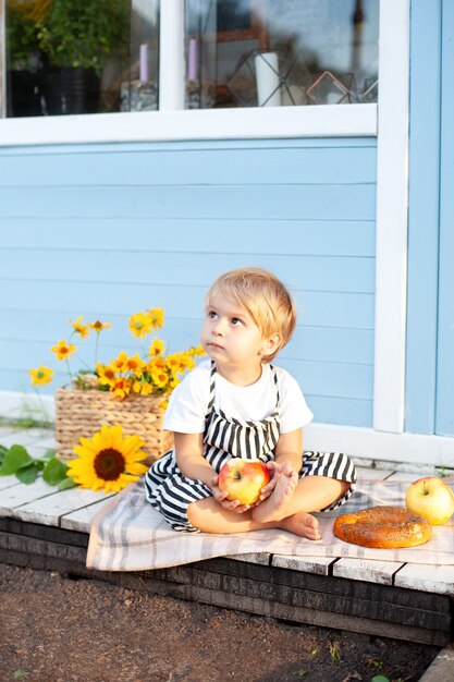 Photo little blond boy sitting on a wooden porch at home and eats an apple on an autumn day. child plays in the yard in the fall.