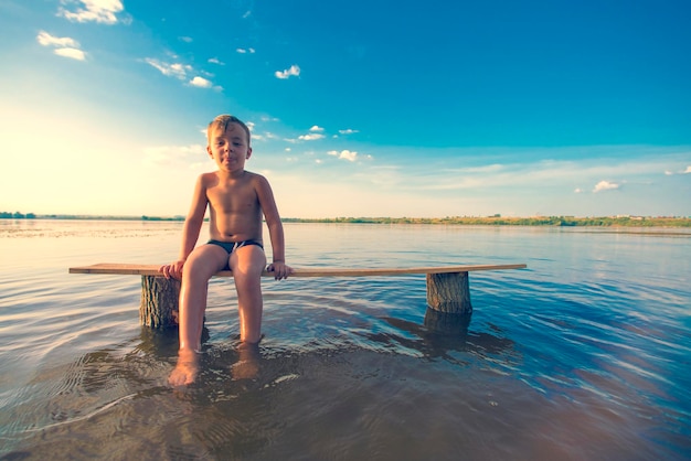 Little blond boy in swimming trunks seated on a wooden bench in the water