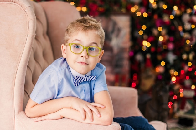 little blonde boy in blue shirt with big glasses sitting on the arnchair