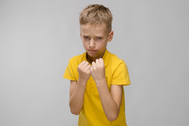 Little blonde caucasian serious sad boy in yellow t-shirt boxing on gray 