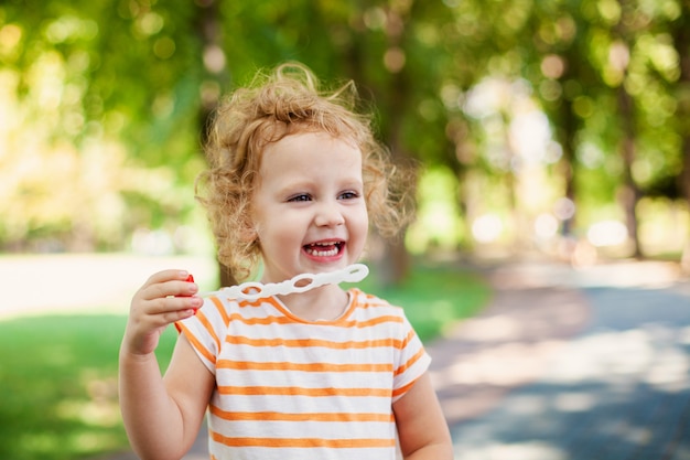 Little blonde curly girl blowing soap bubbles in summer park