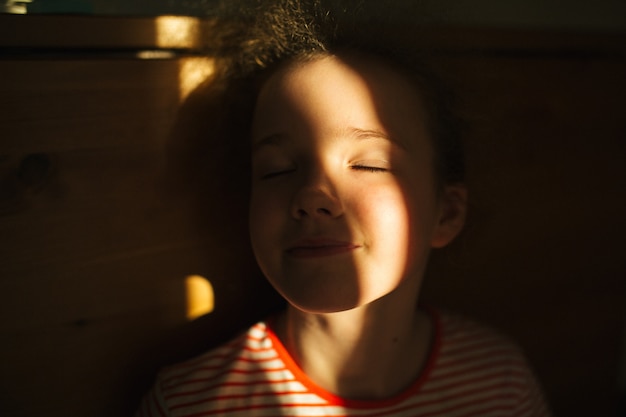 The little blonde cute girl smiles with closed eyes. Sunset warm light. Sunlight in hair. Summer happy time.