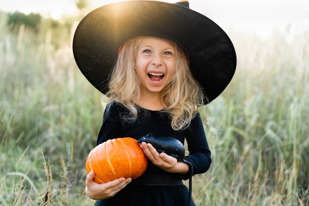 Photo a little blonde girl in a black costume and a witch hat, halloween with a pumpkin and a rat in her hands, a cheerful child.