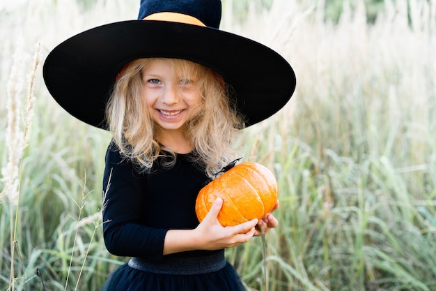 Photo little blonde girl in a black suit and witch hat, halloween with a pumpkin in her hands.