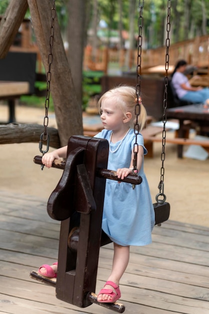 Little blonde girl in blue dress is swinging on wooden swing in summer park outdoors Vertical frame