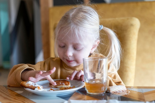 Little blonde girl in cafe at table touches dessert with her hands Child at the table tastes sweets