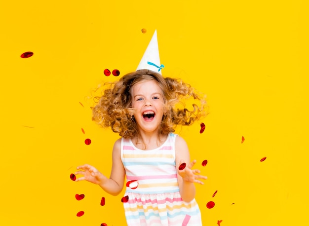 Photo little blonde girl in a festive cap and in a white dress catches confetti a happy smile on a yellow background.