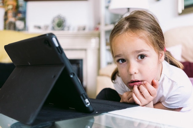 A little blonde girl in front of a tablet looking at the camera in her living room Concept of childhood technology learning having fun the Internet and connectivity