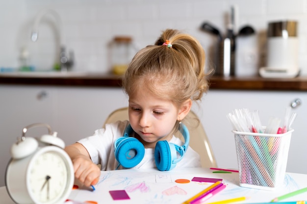 Little blonde girl at home in the kitchen at the table with headphones draws and learns remotely