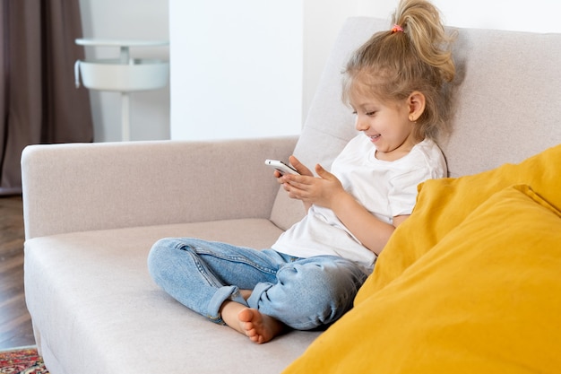 A little blonde girl at home on the sofa with a mobile phone, children and gadgets.