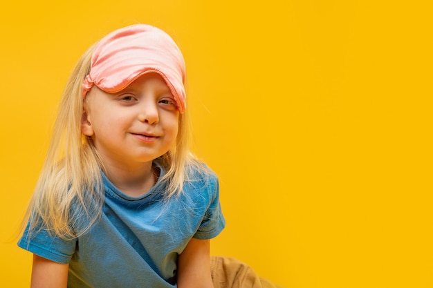 Little blonde girl looks into the camera wearing blue pajamas and pink sleep mask on her forehead Portrait on yellow background