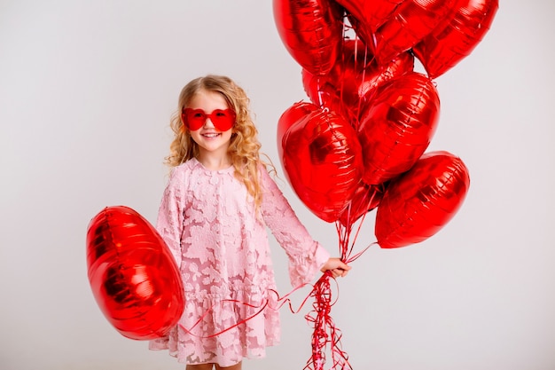 little blonde girl in a pink dress is smiling and holding a lot of red heart-shaped balloons valentines day concept