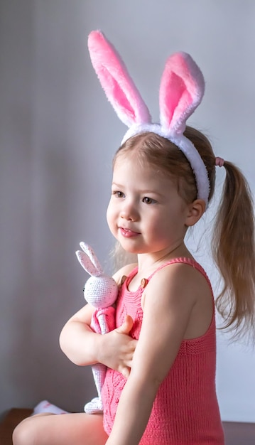 Little blonde girl in a pink romper with the Easter bunny ears on her head hugging a toy bunny