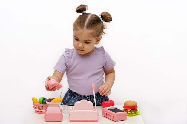 Little blonde girl plays with toy cash register on white table children's vegetable store homemade children's educational games