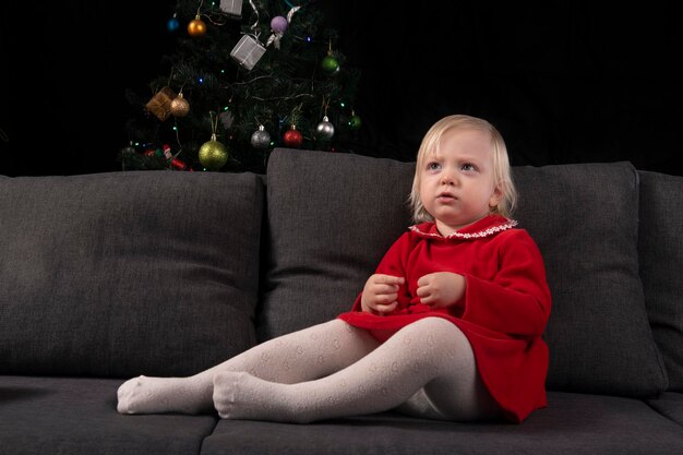 Little blonde girl in a red dress sits on the sofa against the background of a Christmas tree and a black wall. Christmas Eve. New Year