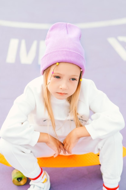 little blonde girl rides a skateboard Stylish girl in a hat sits on a skateboard and poses