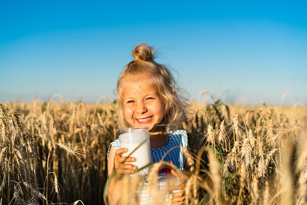 A little blonde girl in a rye field smiles a happy child in the ears with a glass of milk in her