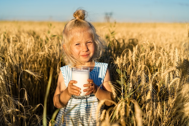 A little blonde girl in a rye field smiles, a happy child in the ears with a glass of milk