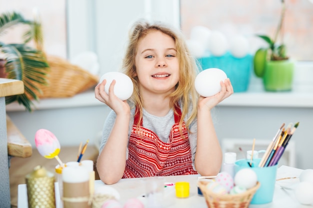 Little blonde girl showing eggs before coloring for Easter holiday at home.