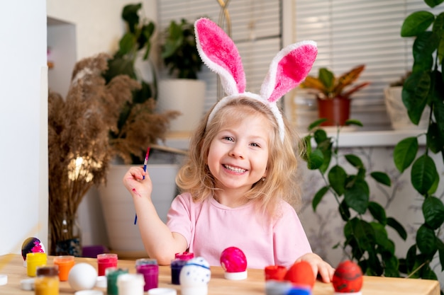 A little blonde girl sits at the table and paints easter eggs with brushes and paints smiles