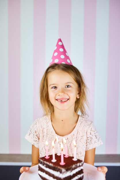 Little blonde girl  smiling  in birthday pink cap, a chocolate birthday cake with candles. Child celebrates her birthday. Happy birthday.