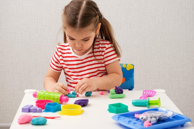 Little blonde girl in striped Tshirt is playing with plasticine at white table at home sculpt different shapes with molds and multicolored modeling clay