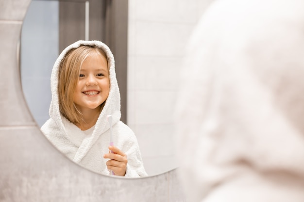 little blonde girl in a white bathrobe brushes her teeth in front of the bathroom mirror