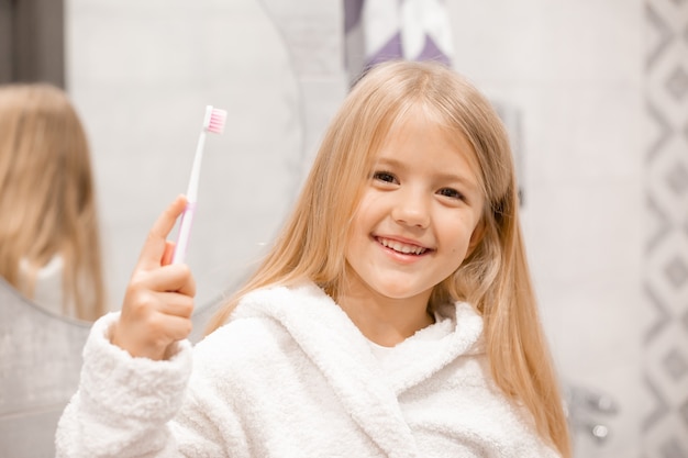 little blonde girl in a white bathrobe brushes her teeth in front of the bathroom mirror