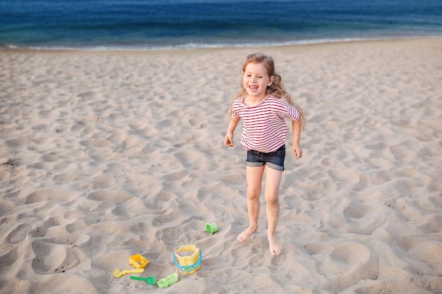 A little blonde girl with long hair in modern clothes plays on the sandy beach and enjoys rest