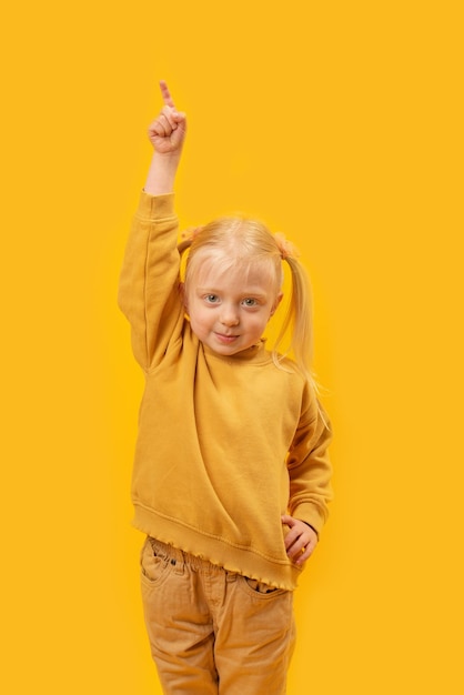 Little blonde preschool girl raised hand up and points index finger at the sky Vertical frame portrait of child in yellow clothes