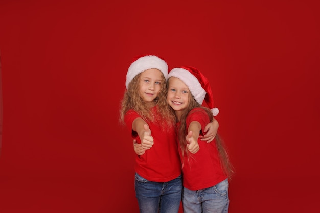 little blonde sisters with blue eyes red tshirts in santa claus hats hug