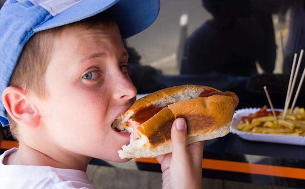 Little boy biting hot dog with ketchup and french fries at fast food restaurant