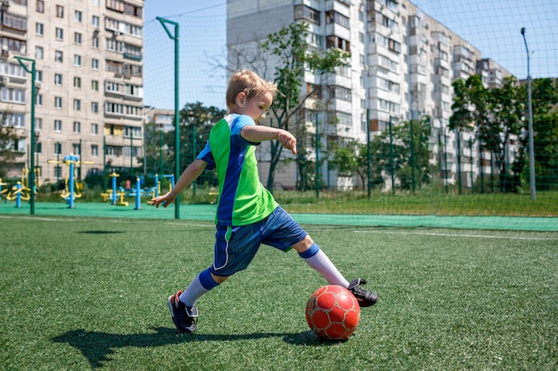 Little boy in blue and green form playing football on open field in the yard a young soccer player