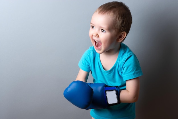 A little boy in boxing gloves and a blue Tshirt on a gray backgroundexpression of emotionscopy space