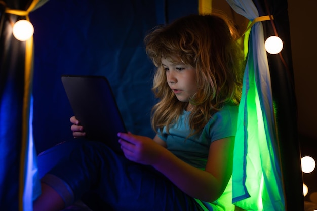 A little boy child uses a laptop at night a child sits in home tent front of a computer screen in th