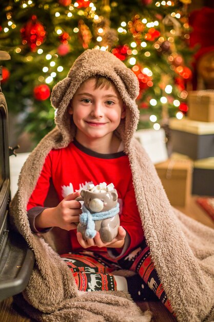 A little boy in Christmas pajamas sits near a Christmas tree