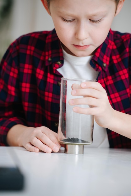 Little boy doing science project, looking at iron filings alligned with a magnet inside a glass. Behind a table.