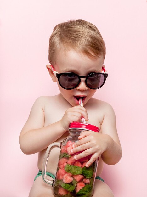 Little boy drinking fresh lemonade with piece of watermelon and ice on pink background