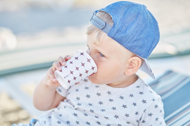 Little boy drinks water from a plastic glass on the beach High quality photo