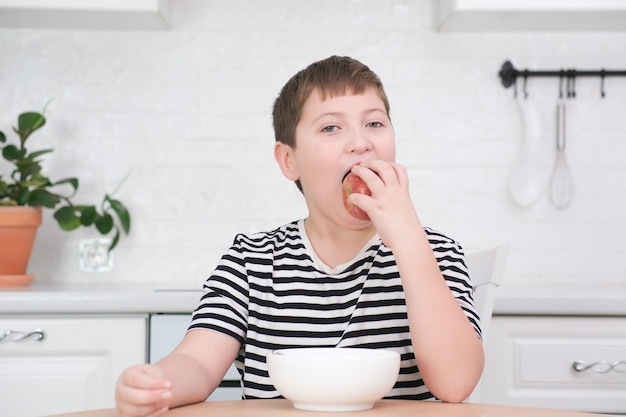 Little boy eating red apple breakfast at wooden table in light white kitchen