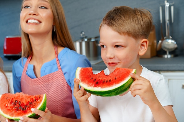 Little boy eating watermelon in kitchen with his mother