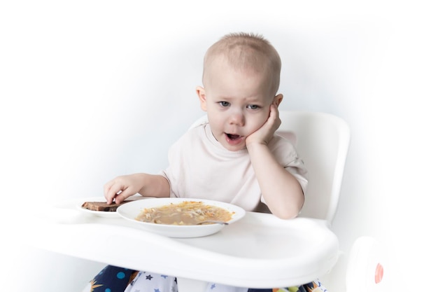 A little boy eats soup with bread on his own in a highchair against the background of a white wall