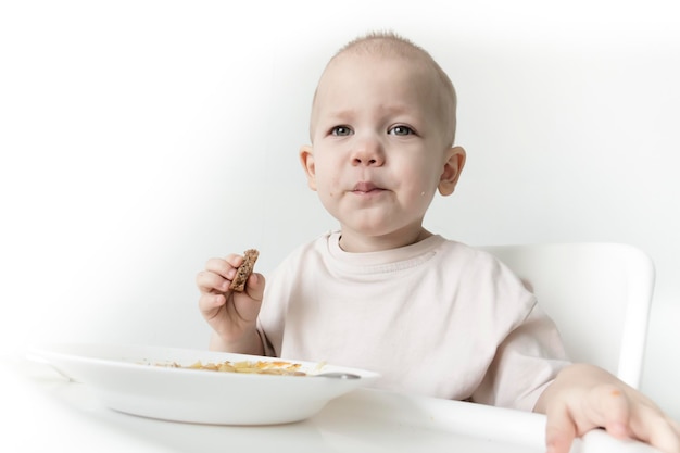 A little boy eats soup with bread on his own in a highchair against the background of a white wall