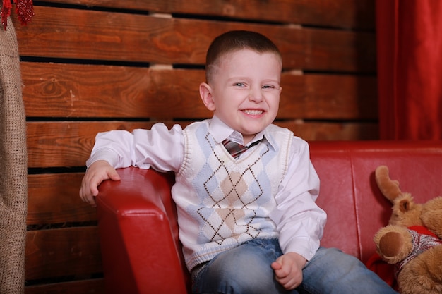 Little boy having fun on the couch in the Christmas studio in red. Christmas tree and teddy bear on the wall.