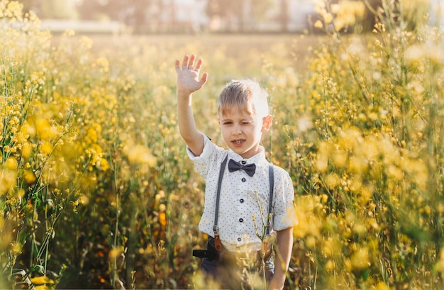 Little boy having fun and waves his hand to the camera in nature in the summer. Cute adorable child in an oilseed rape field, close portrait