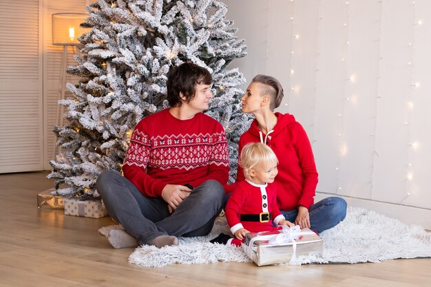 Little boy and his parents at home with Christmas tree on background. Happy boy playing near Christmas tree.